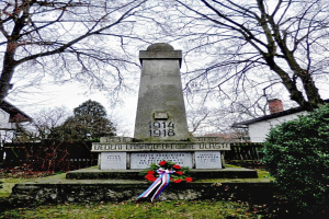 Ein Weltkriegsdenkmal in einem Friedhof, geschmückt mit einem Kränzchen, umgeben von Gras, trockenen Blättern, Pflanzen und Bäumen, mit Häusern und Himmel im Hintergrund.
