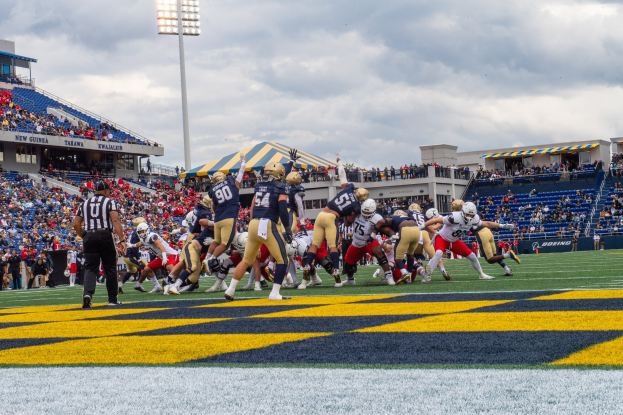 Fußballspieler in Helmen und Sportbekleidung konkurrieren auf einem Feld mit Zuschauern, Stadionstrukturen und Text unten.