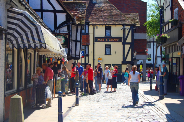 Eine Straße mit Pfählen und Gebäuden auf beiden Seiten, Menschen auf der linken Seite mit einem Handwagen, ein Mann und eine Frau auf der rechten Seite in der Nähe eines lila Müllcontainers, mit Gebäuden, Bäumen und Blumenkübeln im Hintergrund.