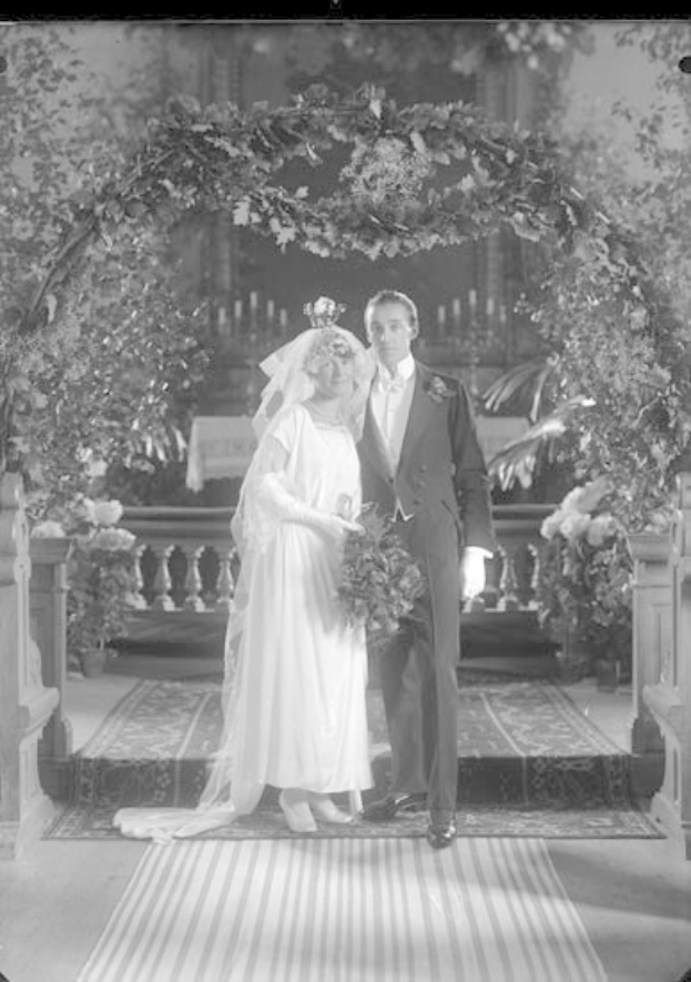 Schwarz-weiß-Foto eines Mannes und einer Frau, die unter einem Blumenschmuckbogen stehen, die Frau hält einen Strauß, mit einem Teppichboden.