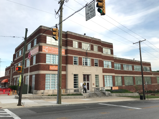 Ein großes Ziegelgebäude an einer Straßenecke mit Strommasten, Ampeln, Schildern, Geländern, Treppen zum Eingang, einer Person auf dem Gehweg, Pflanzen, Gras, Bäumen und einem bewölkten Himmel im Hintergrund, identifiziert als die kürzlich wiedereröffnete Monarchs A-Schule.