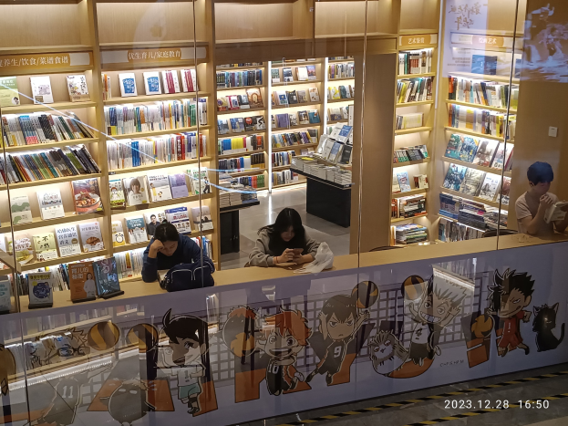 Eine Gruppe von Menschen, die an einem Tresen in einer Bibliothek sitzen, umgeben von Bücherregalen, mit Gegenständen auf dem Tresen und einer Tafel mit Text und Cartoons auf der rechten Seite.