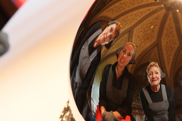 Ein reflektierender Ball auf einem weißen Tisch zeigt das Spiegelbild von drei lachenden Frauen, die sich leicht biegen, mit einem Dach im Hintergrund.