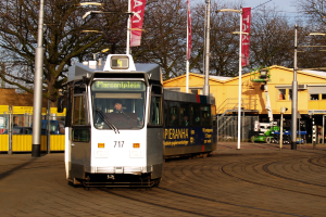 Weiß-blaue Straßenbahn auf einer Stadtstraße mit einem Passagier im Inneren, umgeben von Laternenmasten, Bannern, einem Zaun, anderen Fahrzeugen, Gebäuden, Bäumen und einem klaren blauen Himmel.