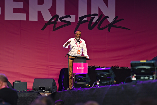 Ein Mann in einem weißen Hemd und braunen Hosen steht auf einer Bühne und hält ein Mikrofon in der Hand, mit einem pinken Banner hinter ihm, auf dem 'Berlin als Fuck' steht und Lautsprecher auf der Bühne.