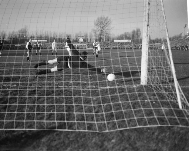 Ein Schwarz-Weiß-Foto eines Fußballspielers, der vor einem Tor einen Ball hält, umgeben von einer Gruppe von Menschen, die auf dem Boden Fußball spielen. Im Hintergrund sind Bäume, Pfosten, Werbetafeln und ein klarer Himmel zu sehen.