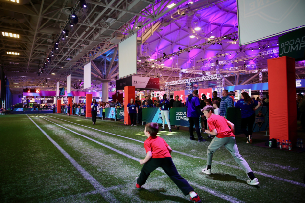 Zwei junge Jungs beim Fußballspielen auf einem Feld mit Zuschauern, umgeben von Flaschen und Gegenständen, mit einem Zaun, Bannern, Anzeigebildschirm, Metallrahmen, Dach, Deckenleuchten und Metallstangen im Hintergrund.