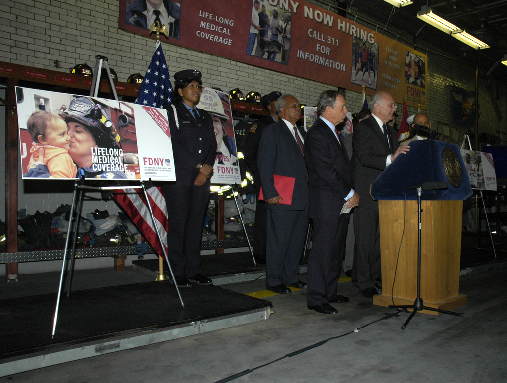 Eine Gruppe von Menschen in der N√§he eines Podiums mit Mikrofonen, Tafeln auf St√§nden, Flaggen und Tafeln an einer Wand, alles unter hellen Lichtern.