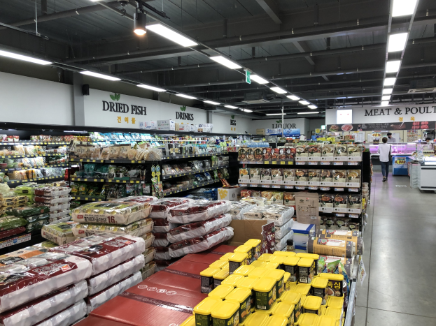 Ein Supermarktgang mit Regalen voller verpackter Lebensmittel, Texttafeln, einer Person, einem Tisch mit Gegenständen und Deckenbeleuchtung.