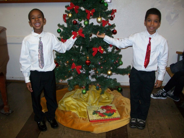 Ein geschmückter Weihnachtsbaum steht in einem Raum mit einem Holztisch links und einem Stuhl rechts, zwei Jungen in weißen Hemden, die strahlen und den Baum halten, eine Schachtel und andere Gegenstände auf dem Boden und eine weiße Wand mit angebrachten Gegenständen im Hintergrund.