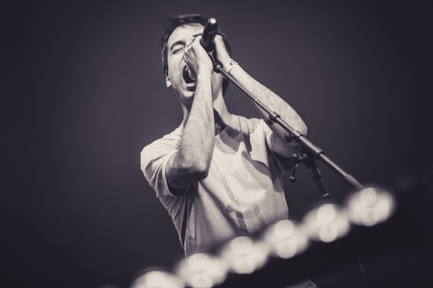 Ein Mann singt in ein Mikrofon auf einer hell erleuchteten Bühne, trägt ein schwarzes T-Shirt und Jeans und hat die Augen geschlossen und eine stylische, aber unordentliche Frisur.