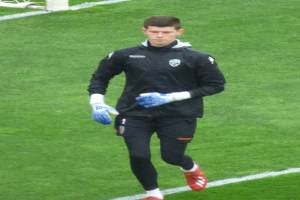 Ein professioneller Fußballspieler in einer schwarzen Jacke und blauen Handschuhen geht auf einem Fußballfeld mit einem Tor im Hintergrund.