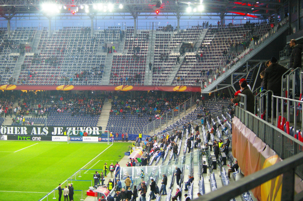 Ein großes Stadion voller Zuschauer bei einem Fußballspiel, mit einigen Personen, die auf dem Feld stehen oder sitzen, einem Geländer und einer Fahne auf der rechten Seite und einem leeren Stadion mit Deckenbeleuchtung im Hintergrund.