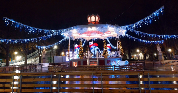 Ein Karussell in einem nächtlichen Park, beleuchtet von Weihnachtslichtern, umgeben von einem Holzzaun, mit Bäumen, Laternenmasten und Fahrzeugen im Hintergrund unter einem dunklen Himmel.