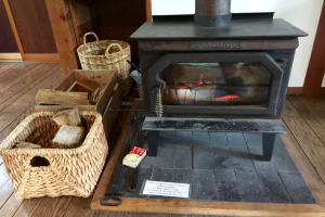 Ein Kaminofen aus Holz auf einem Holzboden neben einem Korb mit Holzscheiten, mit einer Holzbox links und einer Tafel mit Text rechts, vor einer Wand.