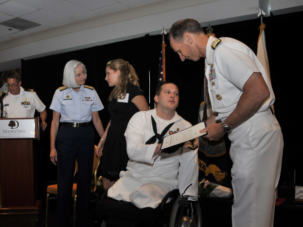 Ein Mann im Rollstuhl erhält eine Auszeichnung von einem stehenden Mann, mit einigen Menschen in der Nähe, einer hält ein Buch, neben einem Podium mit Mikrofon und Texttafel, Fahnen, einem schwarzen Vorhang und einer sichtbaren Decke.