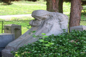 Eine steinerne Statue einer Frau, die auf einem Friedhof sitzt, den Kopf gesenkt und die Hände im Schoß gefaltet, umgeben von Pflanzen, einem Zaun und Bäumen.