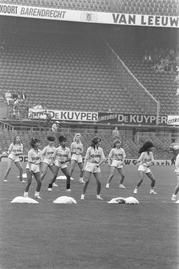 Schwarzes Gruppenbild von Frauen auf einem Fußballfeld mit Zuschauern auf den Rängen und Bannern im Hintergrund.