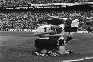 Drei Fußballspieler feiern ein Tor auf dem Boden, umgeben von einer jubelnden Menge in einer schwarz-weißen Stadionszene.