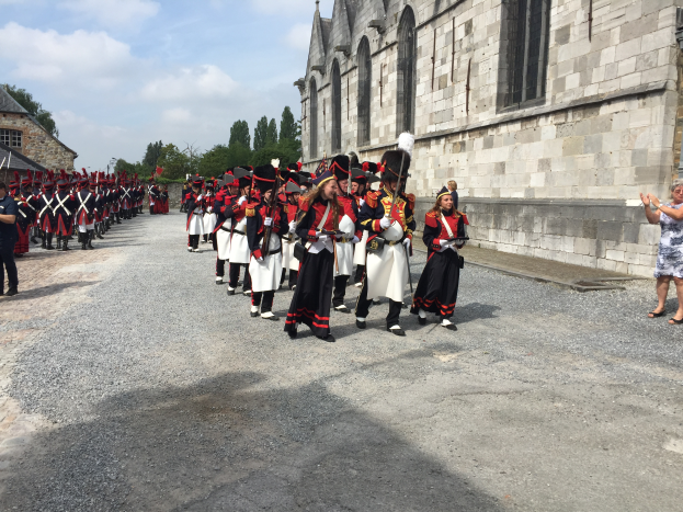 Eine Gruppe uniformierter Personen marschiert auf einer Straße vor einem Gebäude mit Fenstern, umgeben von Bäumen und einem bewölkten Himmel, einige halten Gewehre und andere klatschen.