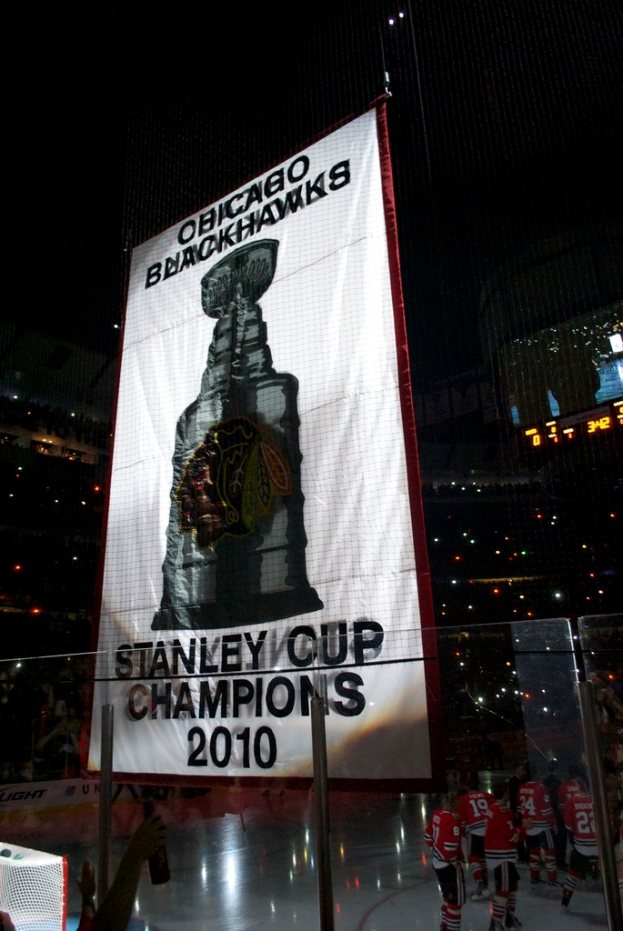 Eine nächtliche Stadion-Szene mit einem großen Banner in der Mitte, Zuschauern im Hintergrund, einem Glas auf dem Boden und einigen Spielern auf dem Feld, beleuchtet von Stadion-Lichtern.
