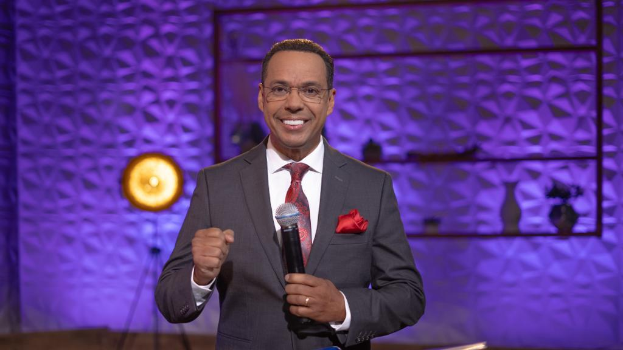 Stephen Colbert, in Anzug, Krawatte und Brille, steht auf einer Bühne und hält ein Mikrofon, mit einem Ständer und einer Lampe dahinter.