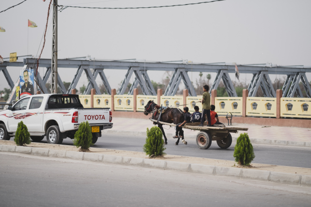 Ein Lastwagen fährt eine Straße entlang neben einem von einem Pferd gezogenen Wagen mit Menschen, mit Pflanzen auf dem Gehweg, einem Mast mit Drähten, Fahnen, einer Brücke, Bäumen und einem klaren blauen Himmel im Hintergrund.