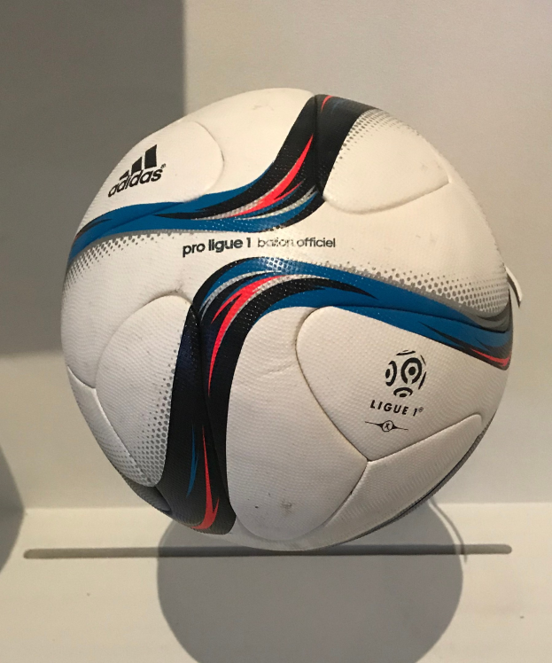 Ein weißer Fußball mit dem Adidas-Logo und der Aufschrift 'Ligue 1' liegt auf einem weißen Tisch gegen einem hellen weißen Hintergrund.