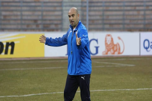 Ein Mann in einer blauen Jacke steht auf einem Fußballfeld während eines Trainings, umgeben von Tafeln mit Text und Logos, und sieht entschlossen aus.