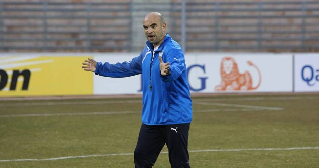 Ein Mann in einer blauen Jacke steht auf einem Fußballfeld während eines Trainings, umgeben von Tafeln mit Text und Logos, und sieht entschlossen aus.