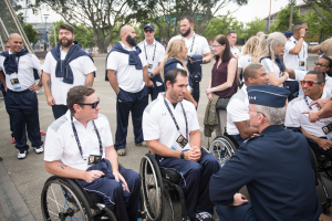 Eine Gruppe von Menschen im Rollstuhl unterhält sich vor einer Kulisse aus Bäumen, Pfählen, Gebäuden und einem klaren blauen Himmel, wobei einige Mützen und Brillen tragen, was darauf hindeutet, dass sie bei den Paralympischen Spielen sind.