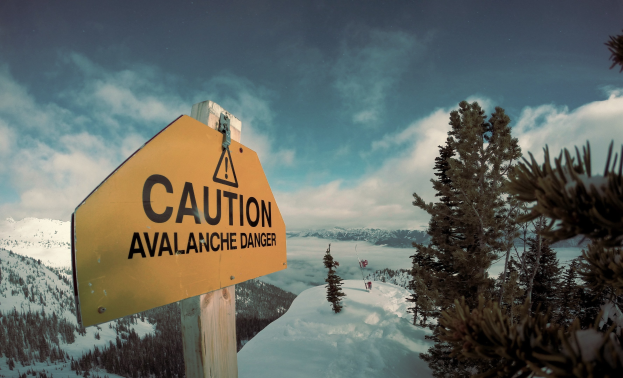 Achtung Lawinengefahr-Schild an einem Holzpfahl in einer schneebedeckten Berglandschaft mit schneebedeckten Bäumen und Hügeln, unter einem sichtbaren Himmel.