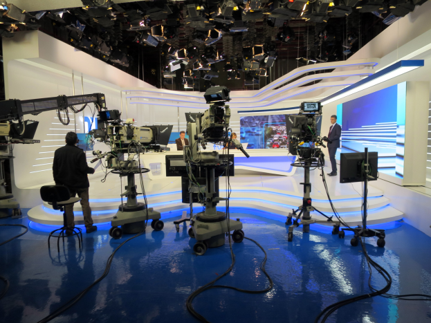 Ein TV-Studio mit mehreren Kameras auf dem Boden, einer sitzenden Person im Vordergrund und einigen stehenden Personen im Hintergrund, mit deckenmontierten Lichtern für eine Live-Broadcast-Aufnahme.