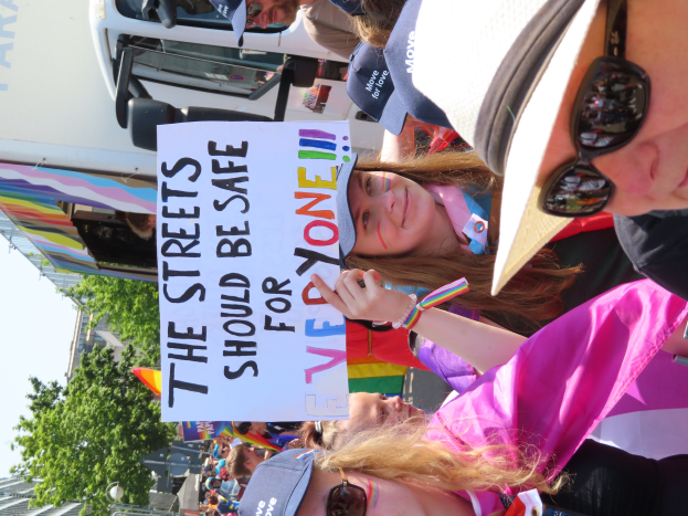 Eine Gruppe von Menschen steht vor einem Lastwagen und hält ein Schild mit der Aufschrift "Die Straßen sollten sicher sein", wobei einige Mützen und Schutzbrillen tragen und andere Schilder halten, vor einem Hintergrund aus Bäumen, Gebäuden und einem klaren blauen Himmel.