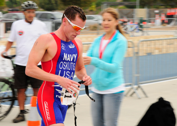 Ein Mann in einem roten und blauen Triathlonanzug läuft eine Straße entlang, neben ihm geht eine Frau, während im Hintergrund Menschen, Fahrräder, Verkehrskegel, Geländer, Fahrzeuge, Bäume und andere Gegenstände zu sehen sind, alles leicht unscharf.