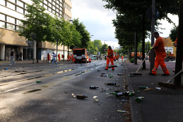 Menschen in orangen Uniformen sammeln Müll von einer Straße mit Schutt, Bäumen und Gebäuden unter einem bewölkten Himmel.