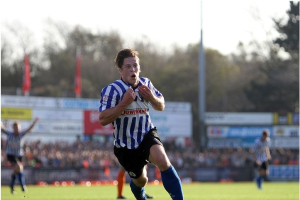 Ein Mann in einem blauen und weißen gestreiften Hemd rennt mit einem Fussball auf einem Rasenfeld während eines Spiels von Huddersfield Town gegen Swansea City, mit Zuschauern, Tafeln, Fahnen und Bäumen im Hintergrund unter einem klaren blauen Himmel.