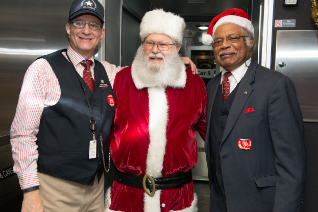 Eine Gruppe von Menschen mit Brillen, Mützen, Krawatten, ID-Karten und Abzeichen, darunter eine Person in einem Weihnachtsoutfit, die vor einer Stahlwand mit Lichtern, Schildern und anderen Gegenständen im Hintergrund steht.