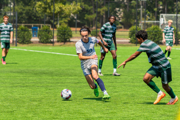 Junge Männer in Sportuniformen spielen Fußball auf einem grünen Rasen mit Bäumen, Maschendrahtzaun und einem Torpfosten.
