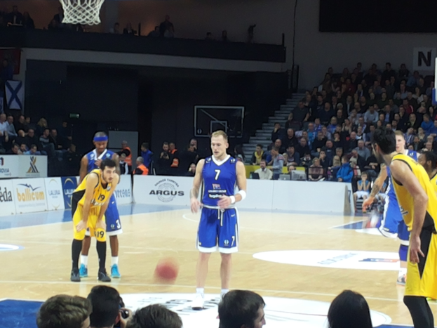 Zwei Mannschaften in blauen und gelben Uniformen spielen Basketball auf einem Indoor-Court während eines 7days Eurocup-Spiels zwischen Fenerbahçe und Real Madrid, mit Zuschauern in den Tribünen hinter ihnen.