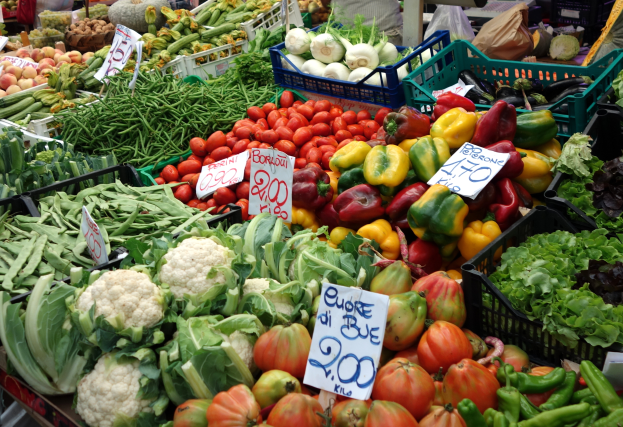 Ein Bauernmarkt mit Körben voller Tomaten, Paprika, Blumenkohl, grünen Bohnen und anderen Gemüsen, Preisetiketten sind sichtbar und einige Menschen in der Nähe.