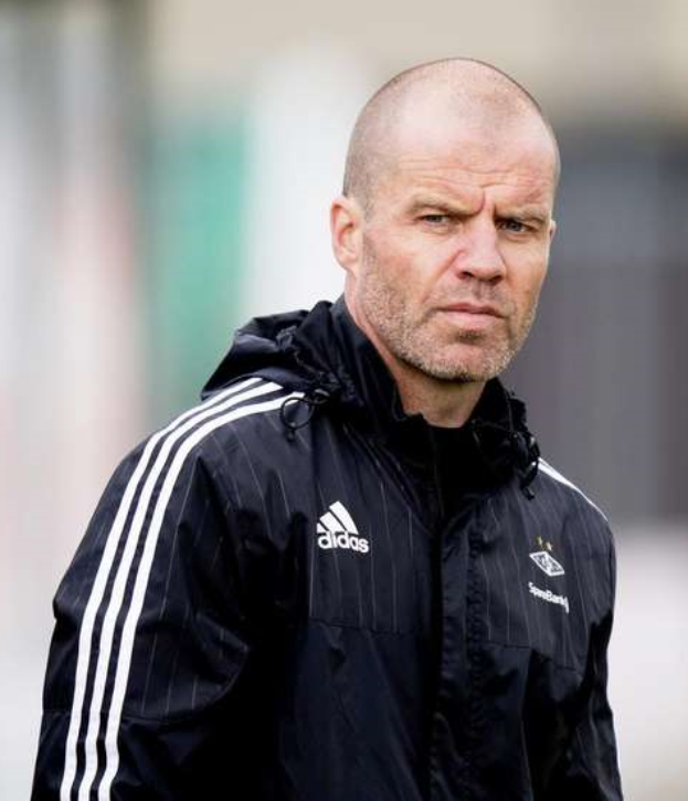 Ein kahlköpfiger Mann in einer schwarzen Jacke steht auf einem Fußballfeld, mit gefalteten Händen und einem ernsten Gesichtsausdruck.