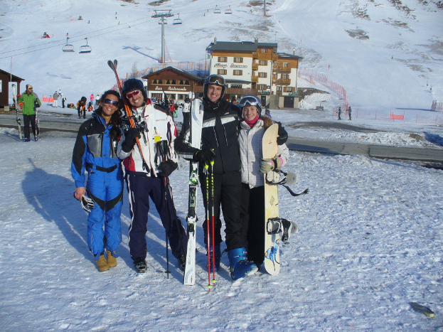 Vier Personen posieren für ein Foto, auf dem sie Skaterpads und Werkzeuge halten, mit Gebäuden und anderen Menschen im Hintergrund, vor einer schneebedeckten und vereisten Berglandschaft.