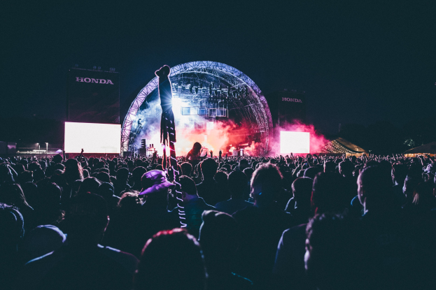 Eine große Menge Menschen auf einem Musikfestival mit einem Mann auf der Bühne, umgeben von Musikern, beleuchtet von Lichtern, Bildschirmen und Bannern vor dunklem Hintergrund.