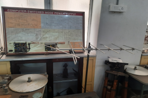 Ein unordentlicher Raum mit verschiedenen Elektronikgeräten, darunter eine beschriftete Leiterplatte auf einem Tisch, Metallstangen, eine wandmontierte Schalttafel, ein Fenster und Gegenstände in Schränken, die auf eine computergeschaltete Modus-Stromversorgung hinweisen.