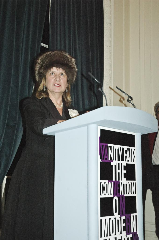 Eine Person steht an einem Podium mit Text darauf und spricht in ein Mikrofon, mit mehreren Personen und Vorhängen im Hintergrund.