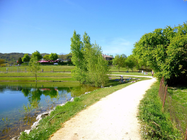 Ein Schotterweg neben einem Gewässer, umgeben von üppiger Vegetation und einem Zaun, mit Menschen, die die Landschaft genießen, Gebäuden, Hügeln und einem klaren blauen Himmel im Hintergrund.