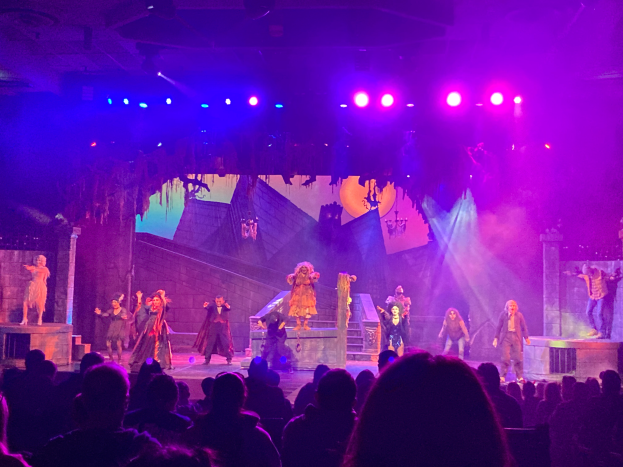 Eine Gruppe von Menschen in Kostümen steht auf einer Bühne unter hellen Scheinwerfern, mit einem Publikum davor und einer Wand im Hintergrund.