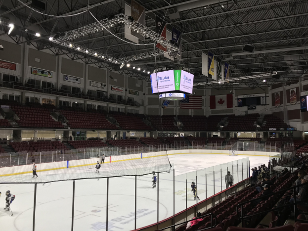 Ein Eishockeyspiel in einer großen Arena mit einem zentralen Tor, Zuschauern auf beiden Seiten, Deckenbeleuchtung und Bannern sowie dem St. Louis Blues Arena im Hintergrund.