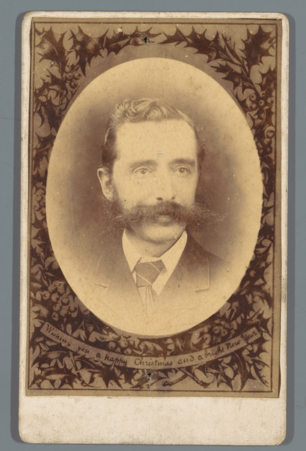 Ein Mann mit Schnurrbart und Bart, der einen Anzug und eine Krawatte trägt, schaut direkt in die Kamera und hat einen ernsten Gesichtsausdruck, seine Hände sind vor der Brust verschränkt.